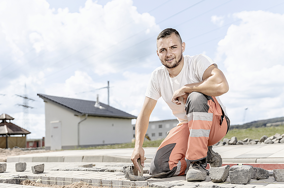 Ein Auszubildender kniet auf einem Bein und lächelt in die Kamera. In seiner linken Hand hält er ein Pflasterstein und in seiner rechten Hand ein Zollstock. 