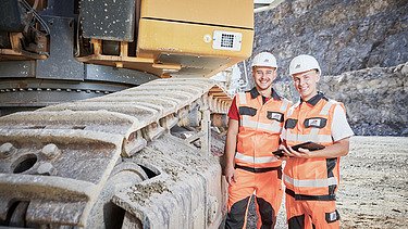 Ausbildung Verfahrensmechaniker:in Zwei Azubis mit Helmen stehen neben einem großen Kettenfahrzeug auf einer Baustelle