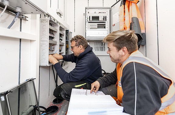 Ausbilder und Auszubildender sitzen in einem kleinen Raum und machen elektrische Installationen..