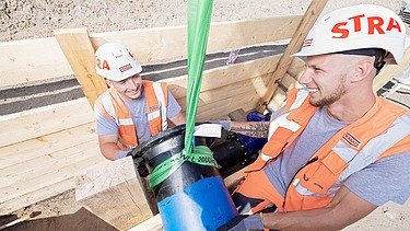 Zwei Auszubildende schieben ein Rohr auf einer Baustelle