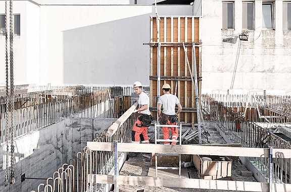 Zwei Hochbaufacharbeiter arbeiten auf einer Baustelle und transportieren einen Holzträger. 