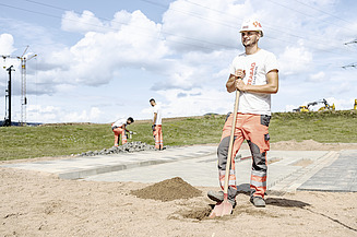 Azubi steht vor Baustelle mit Schaufel in der Hand