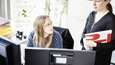 Ausbildung Büromanagement Zwei Frauen stehen vor Bildschirmen und sprechen miteinander