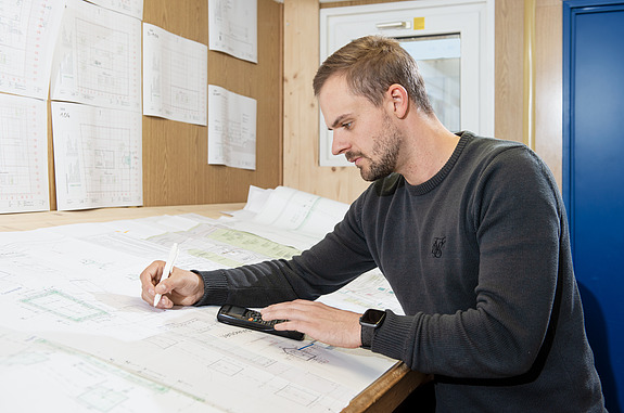 Junger Mann sitzt am Schreibtisch und zeichnet auf einem Bauplan.