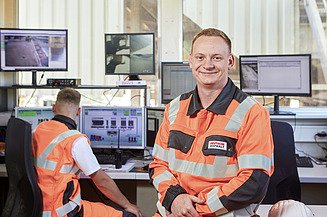 Ausbildung Verfahrensmechaniker:in Mitarbeiter in Arbeitskleidung im Überwachungsraum, einer am Computer, einer schaut in die Kamera
