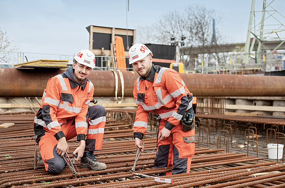 Zwei Auszubildende mit gelben Schutzhelmen schauen nach oben und arbeiten an einem Bauwerk.