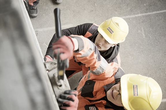 Zwei Auszubildende mit gelben Schutzhelmen schauen nach oben und arbeiten an einem Bauwerk.