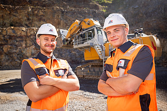 Foto mit 2 Personen, die vor einem Bagger im Steinbruch stehen